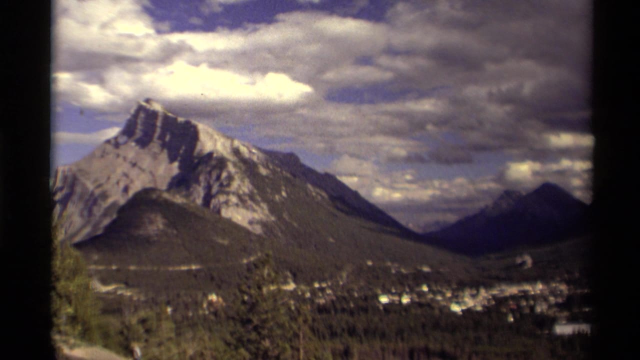 1979 Canadian 8mm view of a snow-streaked mountain, forested valley, small town, and dramatic cloud cover seen through a dark frame edge.