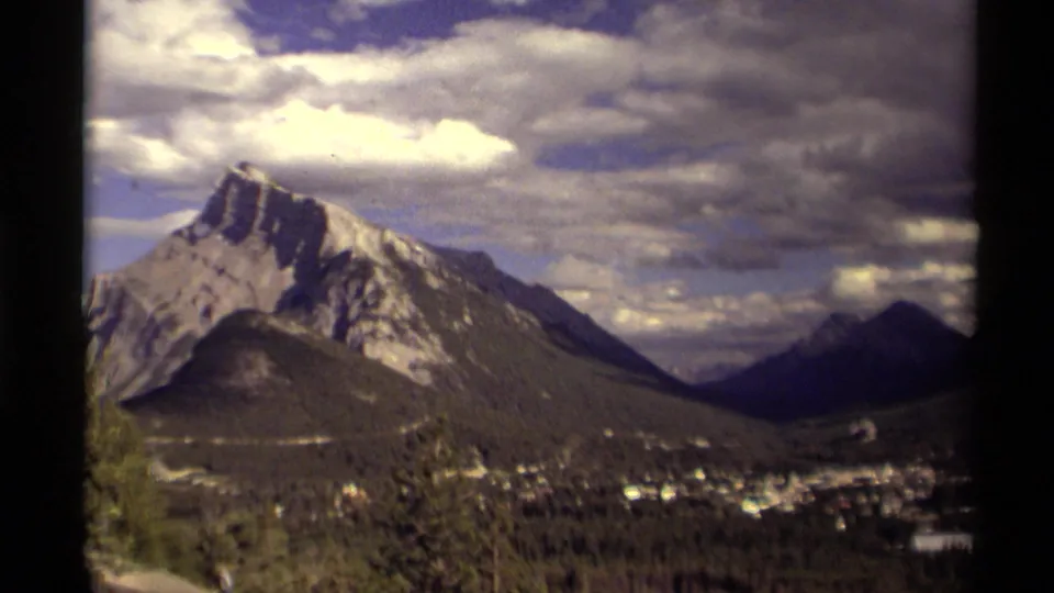 1979 Canadian 8mm view of a snow-streaked mountain, forested valley, small town, and dramatic cloud cover seen through a dark frame edge.