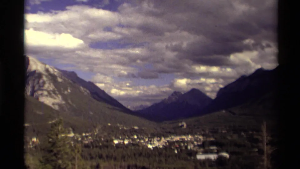 1979 Canadian 8mm view opens wider across a valley town, forested foreground, flanking mountains, distant peak, and layered spring clouds.