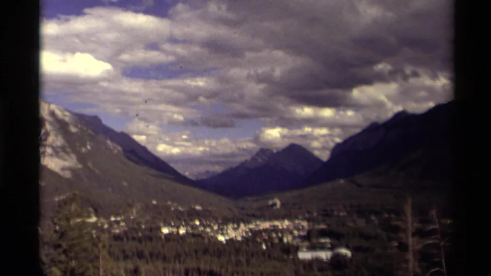 1979 Canadian 8mm frame showing a mountain valley, forest, clustered town, pale distant building, and dark cloud cover from an elevated viewpoint.
