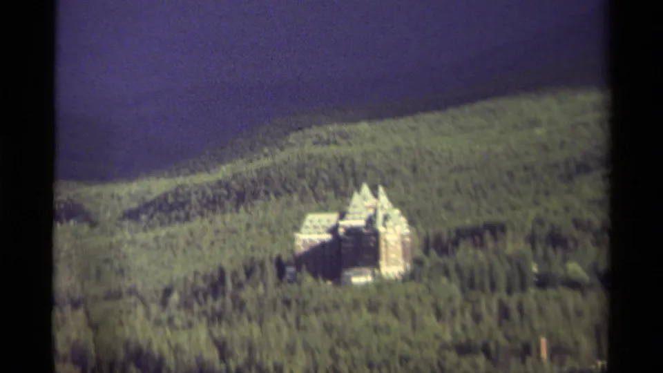 1979 Canadian 8mm frame showing a distant turreted mansion surrounded by dense forest on a hillside under a purple-blue sky.