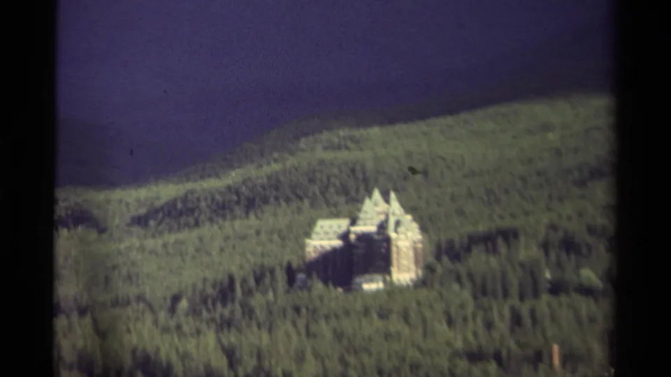 Distant 8mm view of a pale, turreted mansion set within a broad forested slope in Canada, with dark sky above.