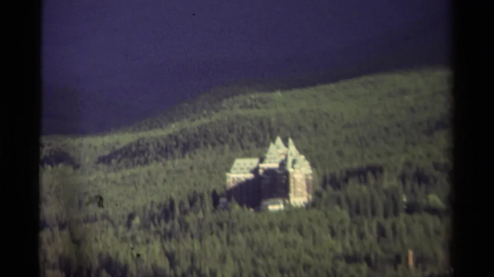 Soft, grainy 1979 Canadian home-movie frame of a castle-like mansion standing among dense trees on a sloped forested hill.