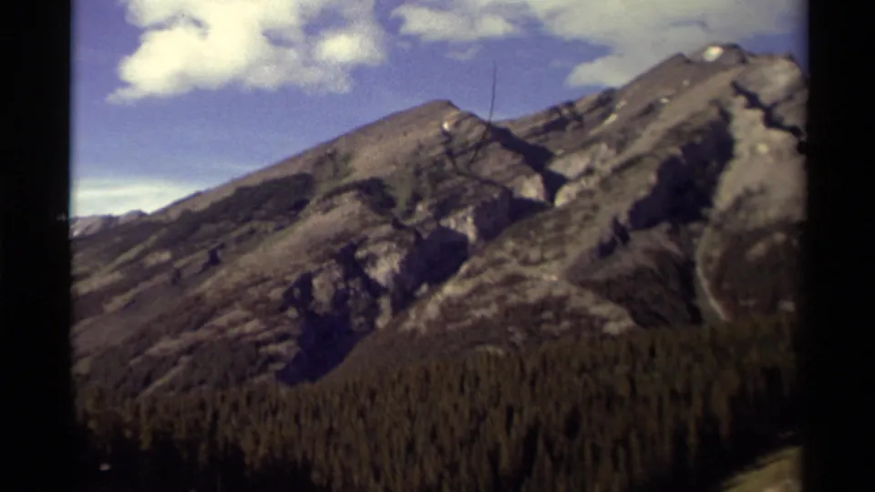 1979 8mm Canada home-movie view of a mountain peak, steep gray slopes, forest, and bright clouded sky.