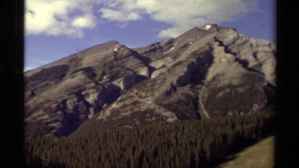 1979 8mm Canada frame showing snow-dusted mountain slopes, forest below, blue sky, and a pale grassy hillside edge.