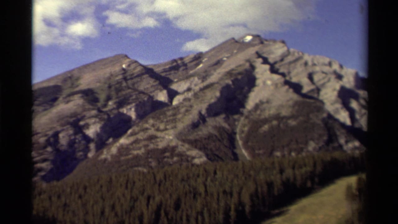 1979 8mm Canada travel footage showing gray mountain peaks, small snow patches, forest below, and blue sky from a moving viewpoint.