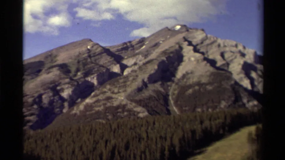 1979 8mm Canada travel footage showing gray mountain peaks, small snow patches, forest below, and blue sky from a moving viewpoint.