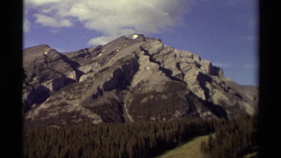 8mm home-movie frame from Canada showing layered mountain rock faces, dark conifer forest, and clouds across a blue sky.