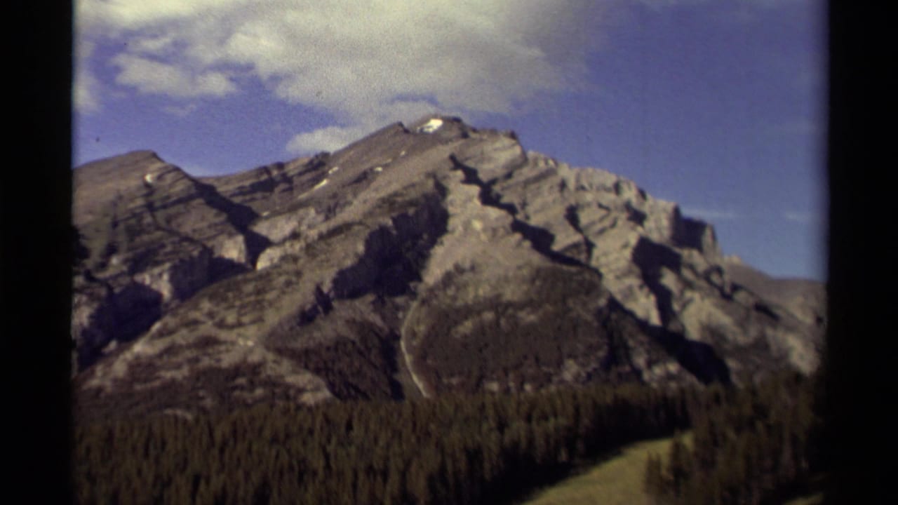 1979 Canadian 8mm frame showing a rocky mountain ridge with a dark evergreen forest in the foreground and pale clouds overhead.