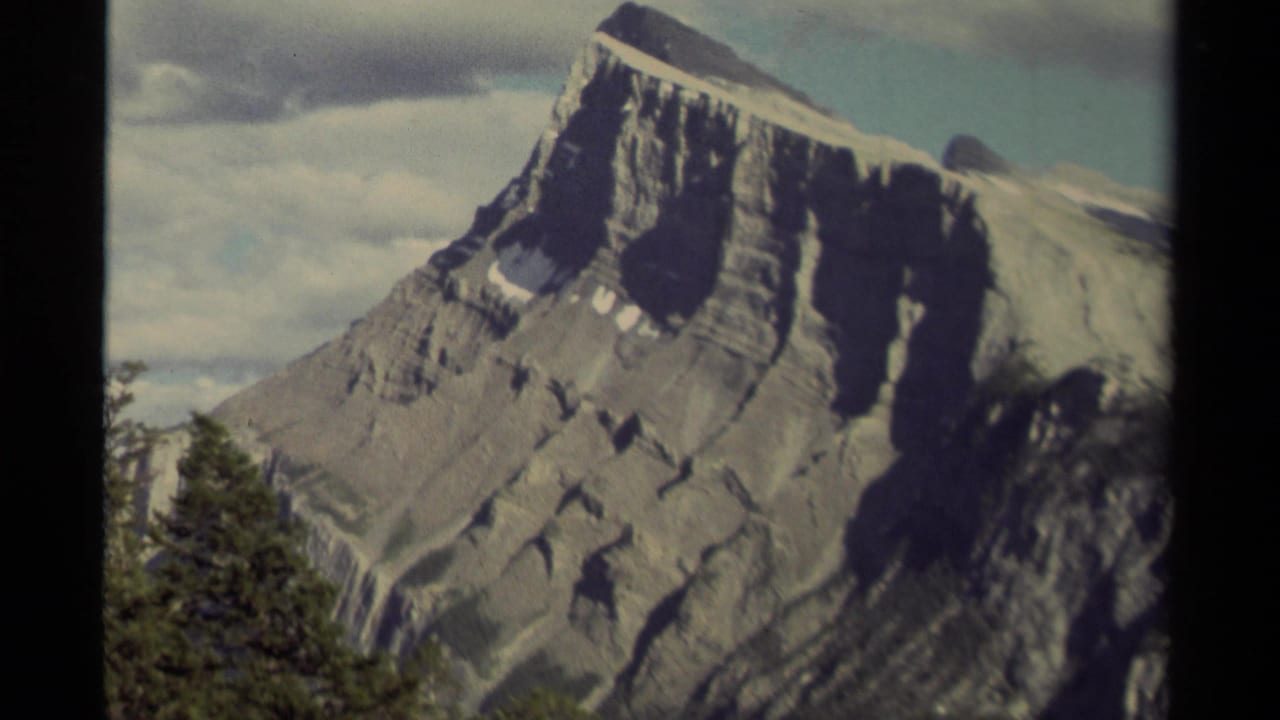 Steep layered mountain face with snow patches, trees, and clouds.