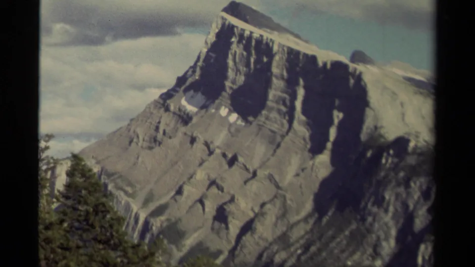 1979 Canadian 8mm frame of a steep layered mountainside with small snow patches, evergreen trees at the base, and cloud cover behind it.