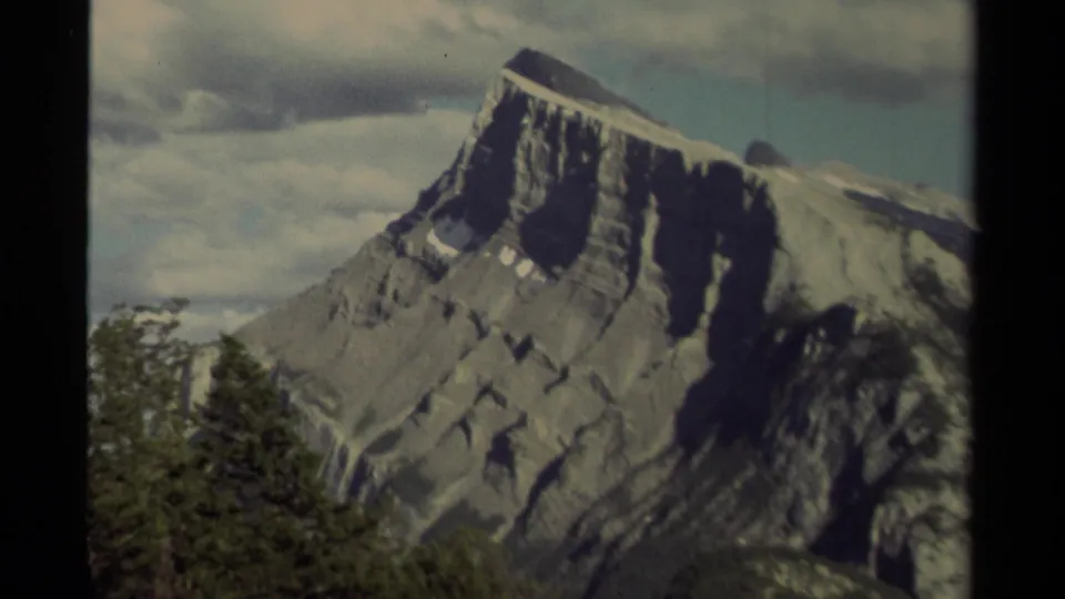 1979 Canadian 8mm frame holding on a large rocky mountain face, with evergreen treetops in front and soft clouds behind the peak.