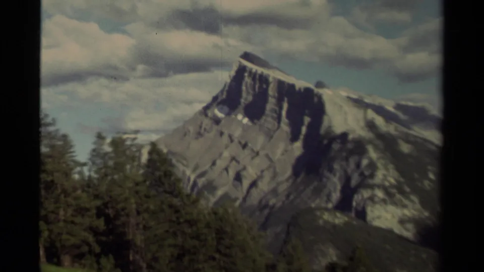 1979 Canadian 8mm frame showing the mountain slope partly framed by dark film borders, with dense evergreens below and clouds across the sky.