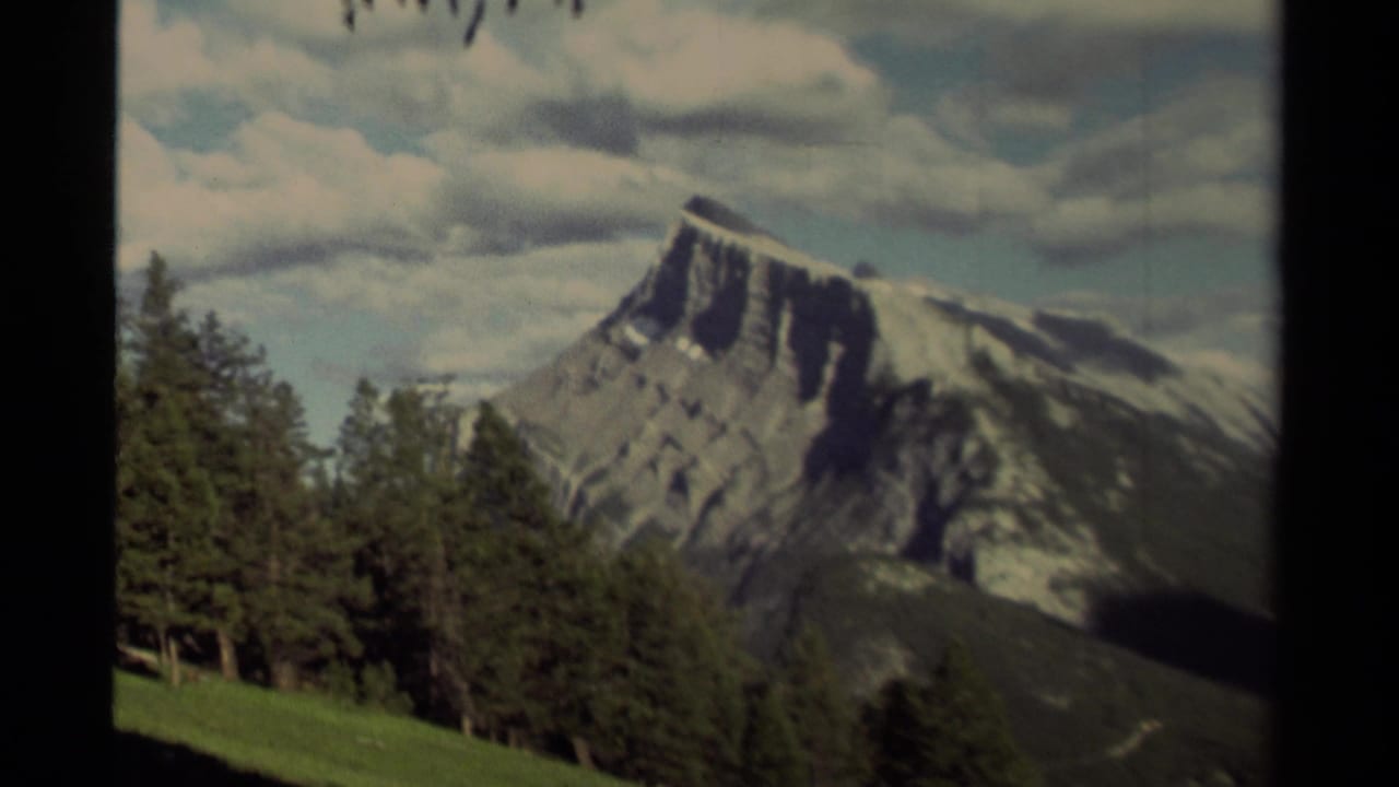 1979 Canadian 8mm view of a snow-streaked mountain peak framed by conifers and a green slope under a partly cloudy sky.