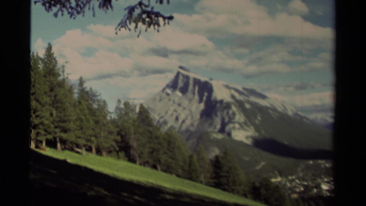 Mountain peak, conifer stand, green hillside, and clouded sky seen through film borders.