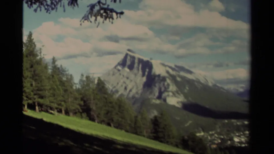 A 1979 8mm Canada mountain view shows foreground pines and a descending green slope leading toward a snow-marked peak.