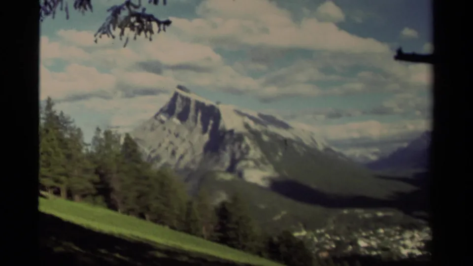 Late-1970s 8mm view in Canada of a mountain peak over a shadowed valley, with pines and clouds framing the scene.