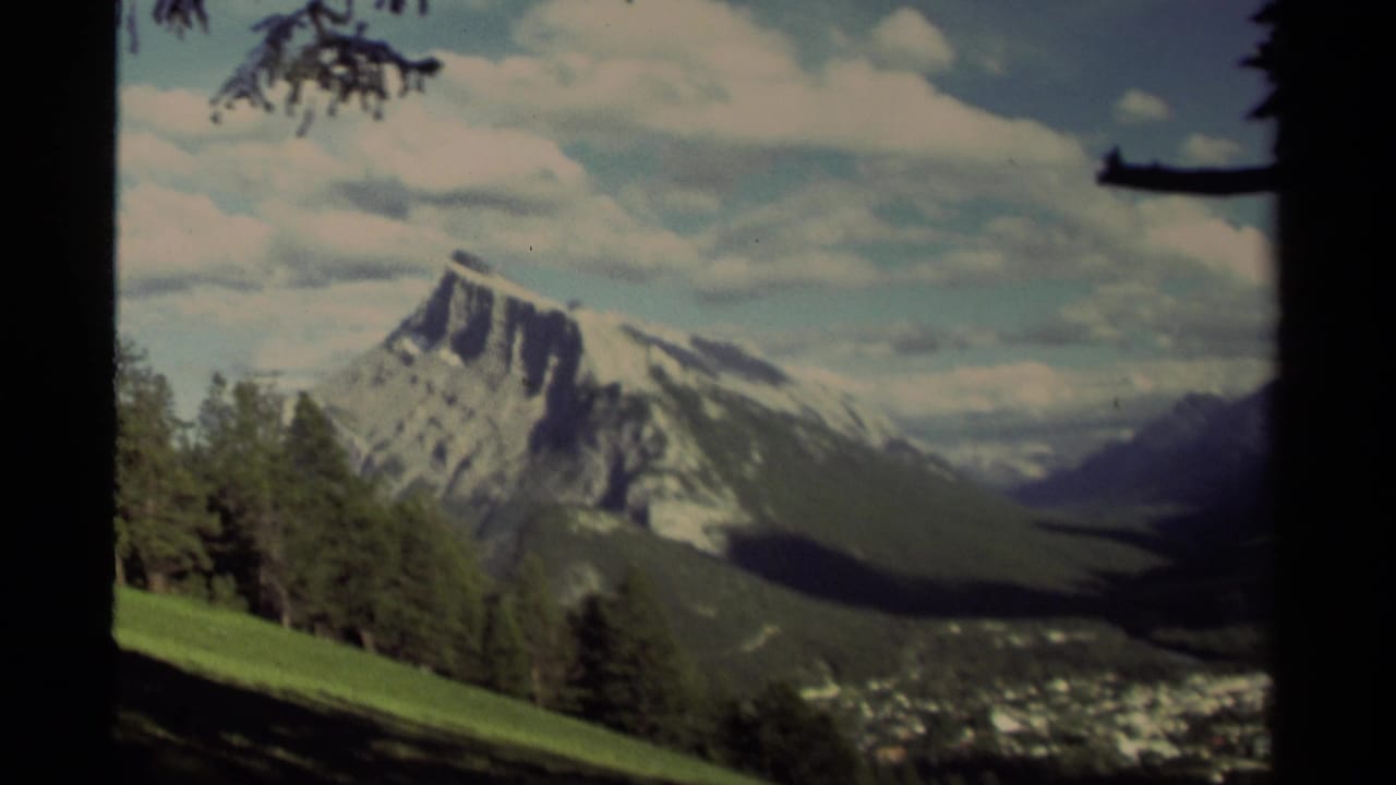 Snow-capped mountain above a green valley, framed by evergreen trees and dark film edges.