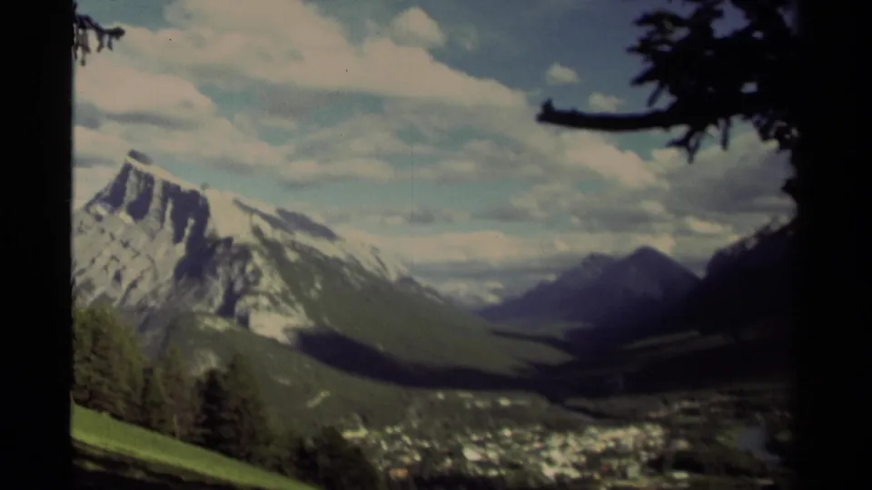 1979 Canadian 8mm view panning across a mountain valley, with the peak partly at left, town below, green slope, and dark tree framing.