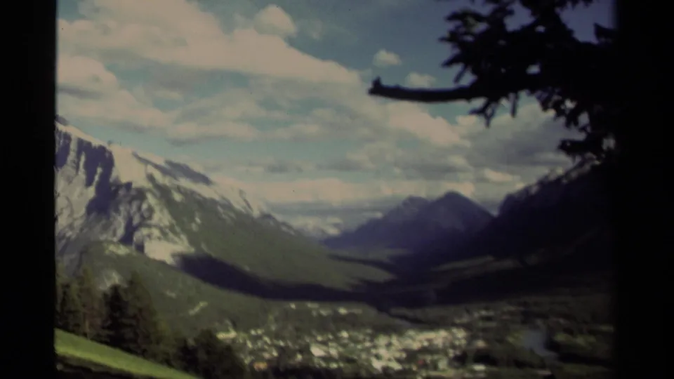 1979 Canadian landscape frame with a shadowed foreground tree, valley floor, distant mountain ridge, green hillside, and bright clouded sky.