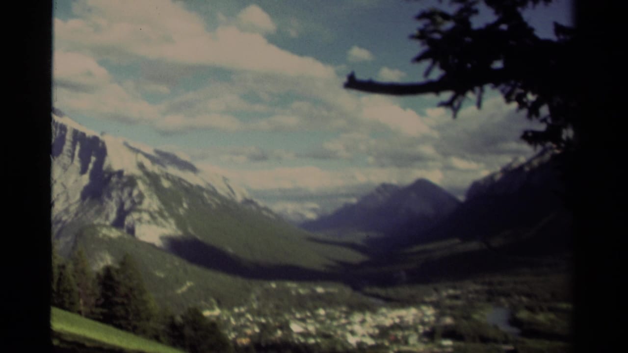 1979 Canadian 8mm frame of a sunlit mountain valley, snow-streaked peaks, a small settlement below, and dark foreground branches.
