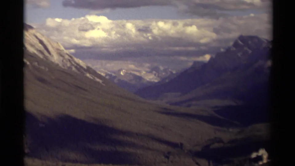 1979 Canadian 8mm frame showing a broad mountain valley, snow-streaked ridges, bright cloud cover, and a small structure near the bottom right.