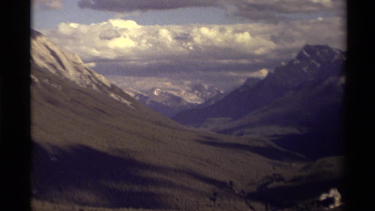 Wide mountain valley view with cloudy sky, rugged slopes, and dark film border.