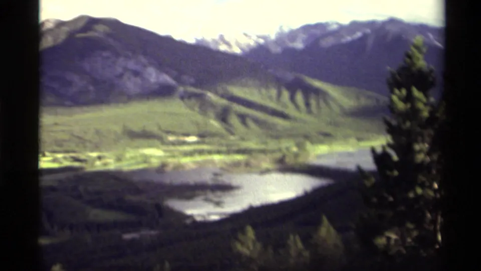1979 Canadian 8mm frame showing a lake in a mountain valley, distant ridges, pale high peaks, and an evergreen tree at the right edge.