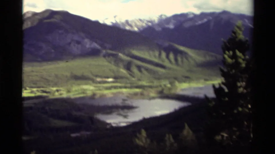 1979 Canadian 8mm frame of a broad mountain-and-lake landscape, with evergreen branches at right and soft, grainy home-movie color.