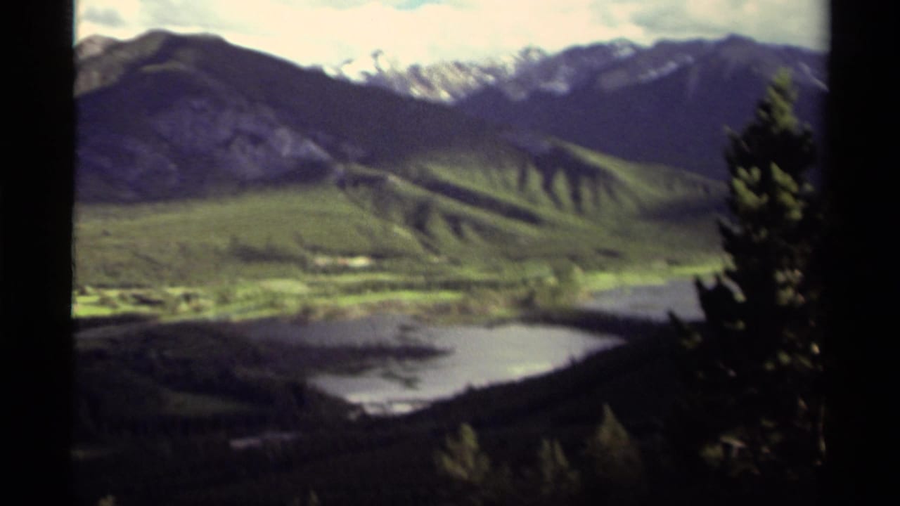 1979 Canadian 8mm frame showing a soft-focus mountain valley with water below, green slopes, snow-touched peaks, and trees along the edge.