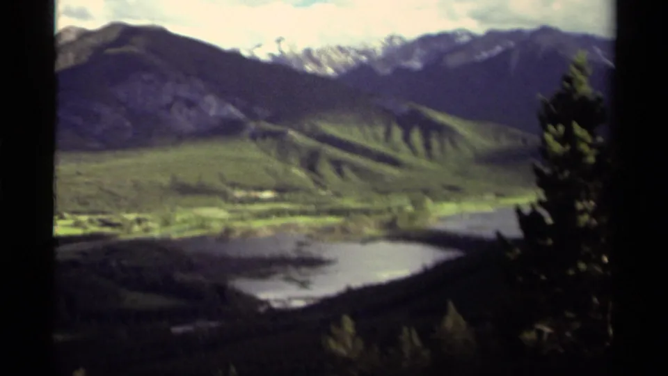 1979 Canadian 8mm frame showing a soft-focus mountain valley with water below, green slopes, snow-touched peaks, and trees along the edge.