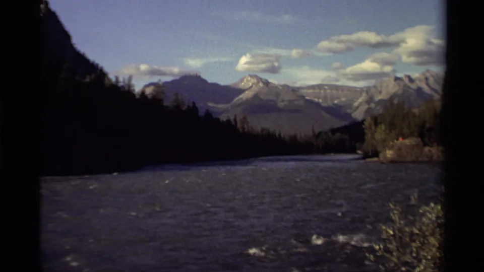 1979 Canadian 8mm frame showing a flowing river, evergreen forest, rugged mountain peaks, and scattered clouds under blue sky.
