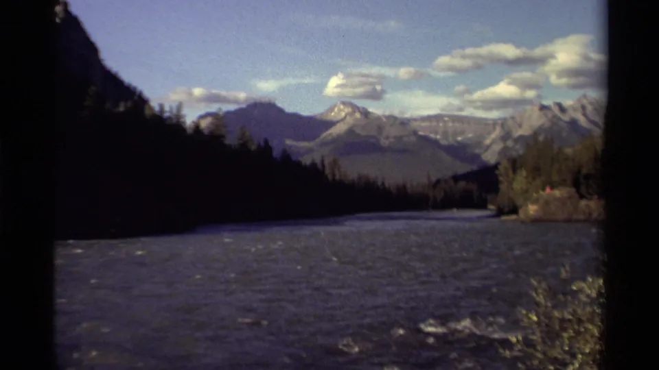 1979 Canadian 8mm frame showing a broad river, dark evergreen slopes, and distant mountain peaks beneath a bright clouded sky.