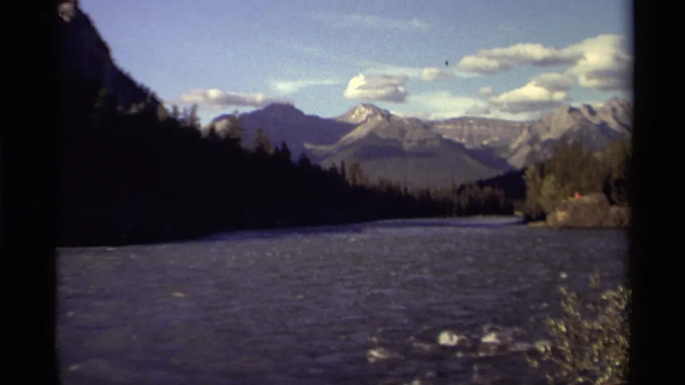 Canadian home-movie frame from 1979 showing a river valley bordered by evergreens with mountain peaks and clouds beyond.