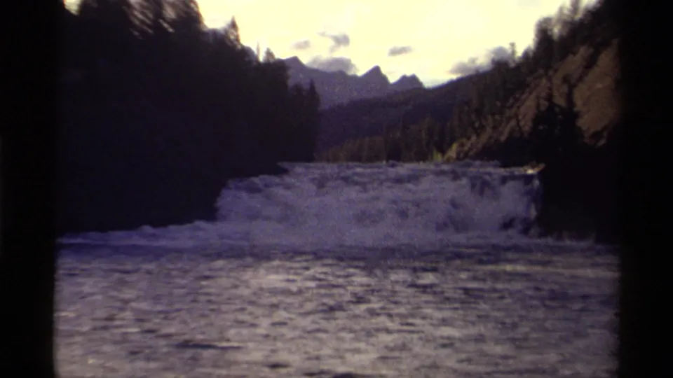The camera view widens to show Canadian rapids, dark river water, tree-lined banks, distant mountains, and a bright cloudy sky.
