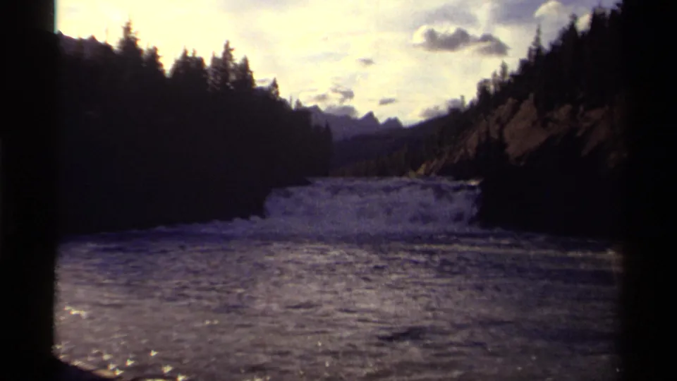 Wide 1979 8mm view of Canadian whitewater flowing below dark forested hills and distant mountain silhouettes.