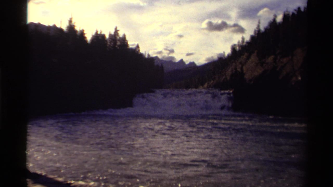 1979 Canadian 8mm frame showing a river flowing between shadowed evergreen slopes with distant mountains and cloud cover.