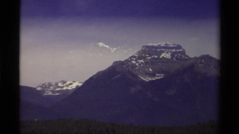 1979 Canadian 8mm frame showing a snow-dusted mountain peak, dark forest foreground, and purple-gray overcast sky.