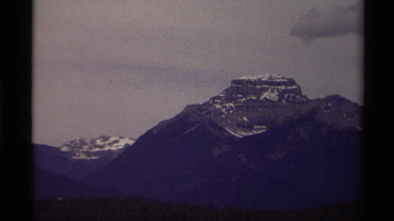 1979 Canadian 8mm frame of a rugged mountain peak with snow patches, dark ridges, and a cloudy muted sky.