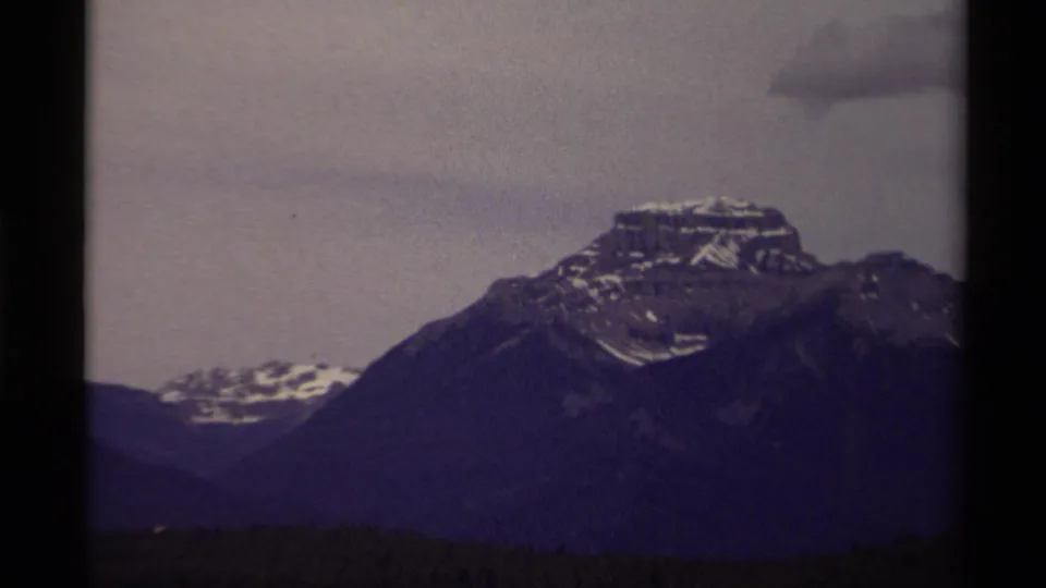 1979 Canadian 8mm frame of a rugged mountain peak with snow patches, dark ridges, and a cloudy muted sky.