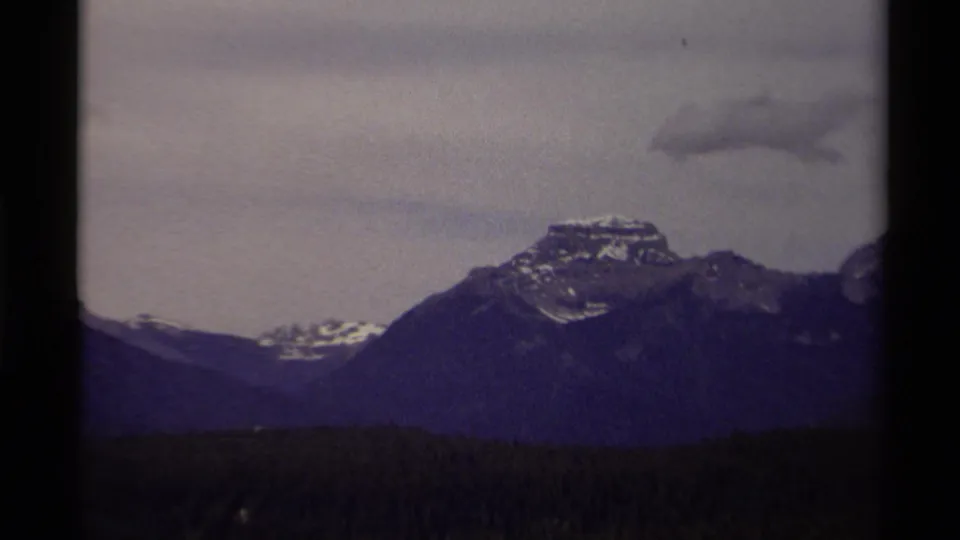 1979 Canadian 8mm frame showing a broader mountain ridge, snow traces, dark trees, and a low overcast sky.