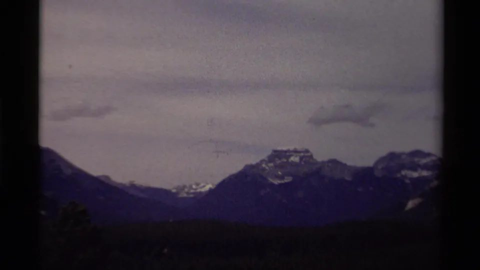1979 Canadian 8mm frame showing a wide mountain range, forested foreground, snow-dusted peaks, and low gray clouds.
