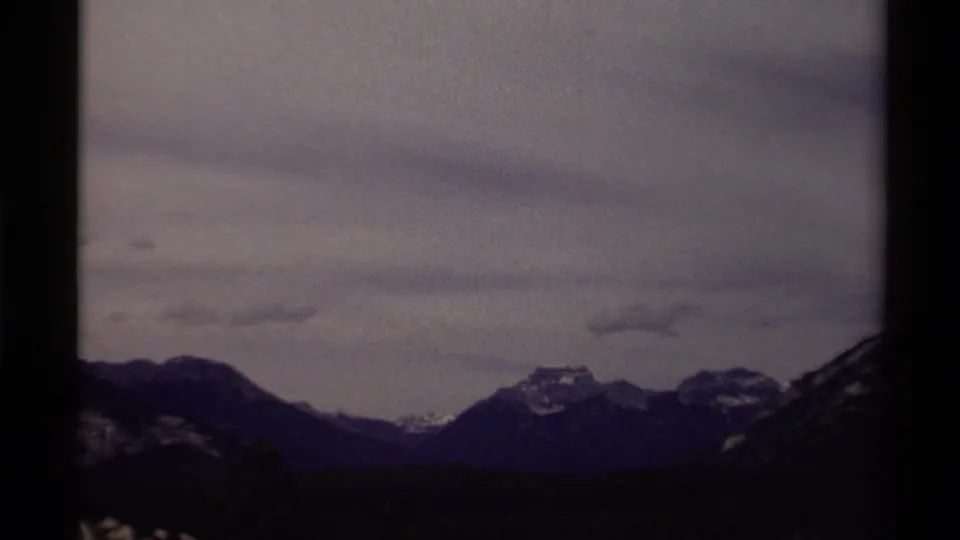 Canadian 8mm travel frame with a distant ridge, snow-dusted peaks, dark forest line, and overcast sky.