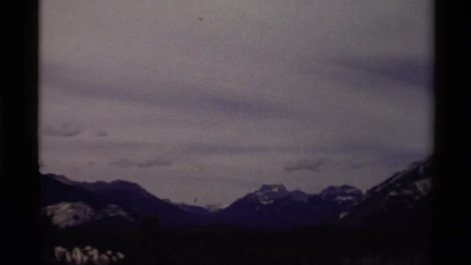 1979 8mm frame from Canada showing low forest, distant snowy peaks, and a pale overcast sky.