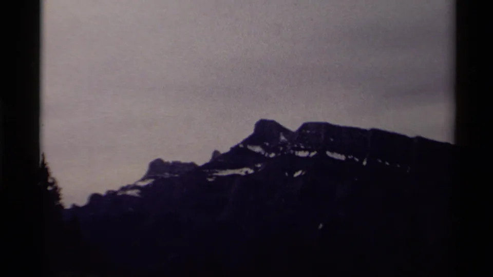 Archival 8mm frame from 1979 Canada showing a low dark mountain range, scattered snow, cloudy sky, and silhouetted foreground edges.