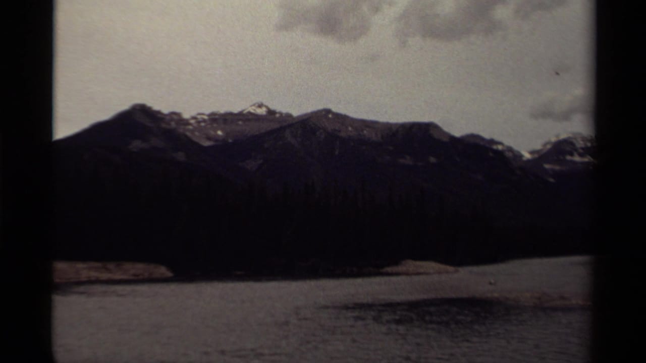 1979 Canadian 8mm frame showing a mountain ridge and calm lake, with snow traces on the peaks and an overcast sky.