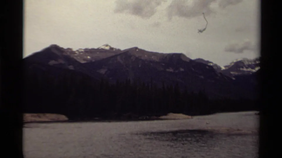 1979 Canadian 8mm frame of a mountain lake bordered by dark forest, with snow-dusted peaks under a pale cloudy sky.