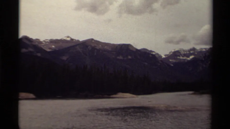 1979 Canadian 8mm frame showing a wide mountain lake scene with forested shore, snow-dusted peaks, and muted overcast light.