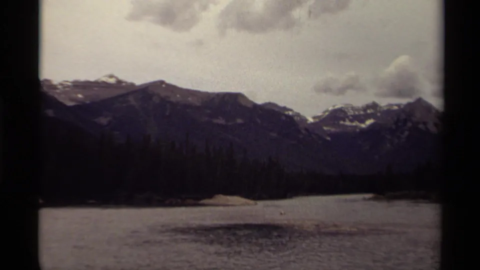 1979 Canadian 8mm frame showing snow-dusted mountain peaks beyond a lake and dark evergreen shoreline.