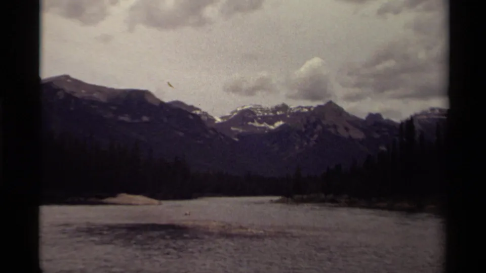 1979 8mm Canadian landscape frame with water in the foreground, trees along shore, and snow-streaked peaks beyond.